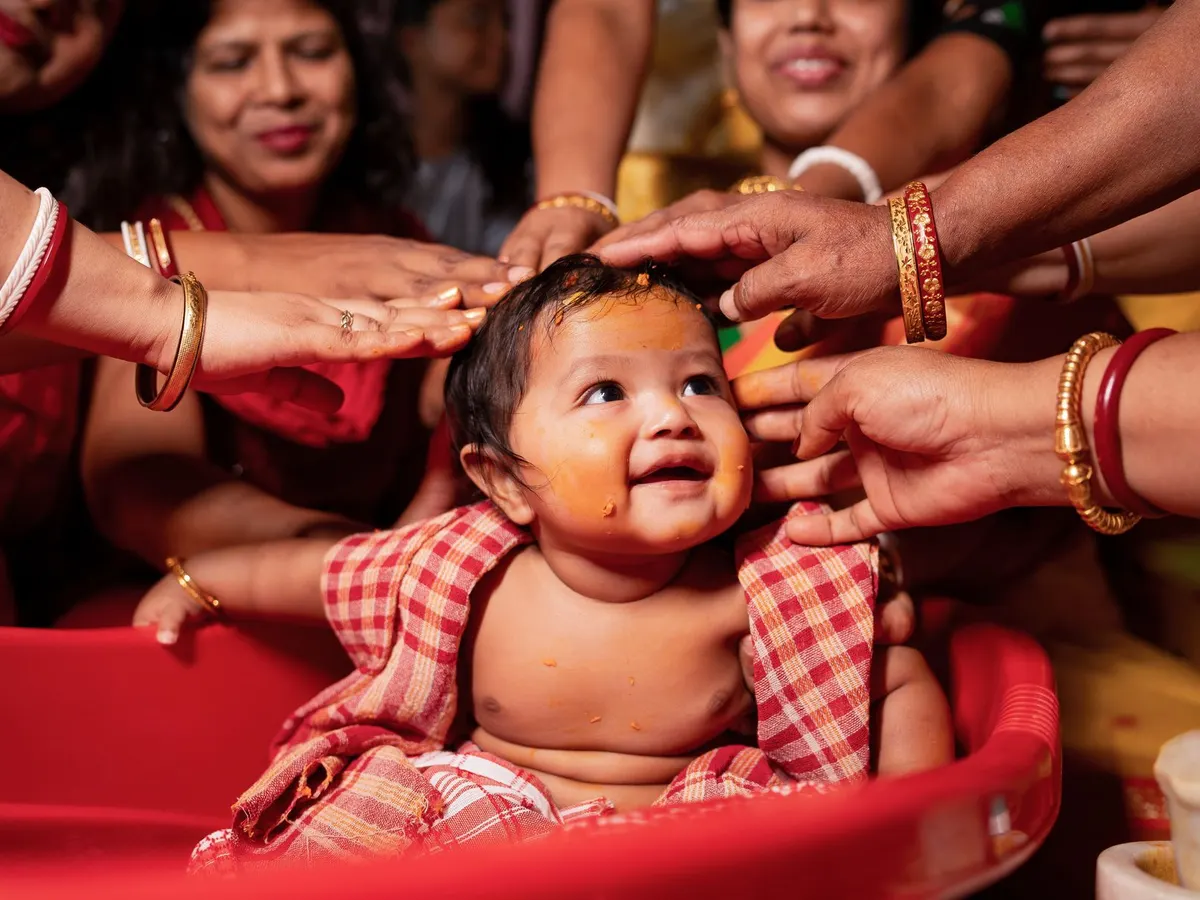 Smiling baby with turmeric on his face, blessed by many hands in red and gold bangles
