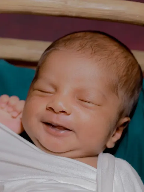 Newborn baby smiling with eyes closed, swaddled in white in a wooden cradle