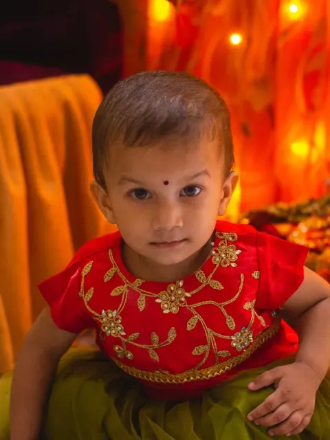 Toddler with a bindi in a red embroidered outfit, festive lights behind